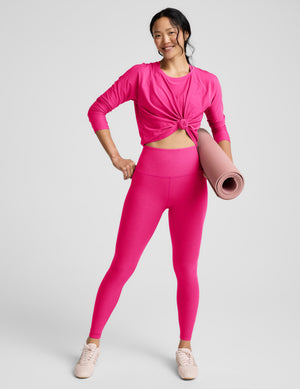 Woman wearing a bright pink long-sleeve top and leggings and holding a yoga mat on a light gray background. 