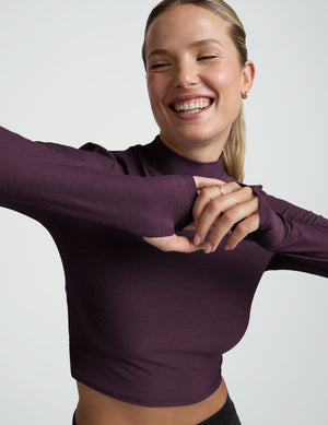 woman wearing a cropped purple long-sleeve mock-neck top and black pants on a plain background. 