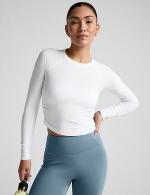 Woman wearing a white long-sleeve top and blue leggings against a gray background. 