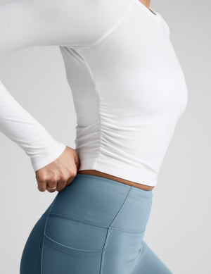 Woman wearing a white long-sleeve top and blue leggings against a gray background. 