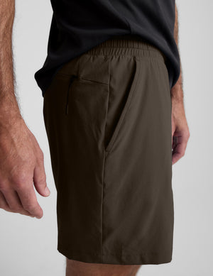 A close-up of a person wearing dark olive brown athletic shorts with side pockets and an elastic waistband, paired with a black t-shirt against a light gray background.