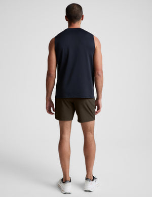 A man viewed from behind wearing a sleeveless navy athletic tank top, dark olive shorts, and white sneakers against a light gray studio background.