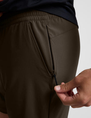 Close-up of a person wearing dark olive brown athletic shorts, showing a side zip pocket being opened by a hand.