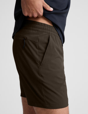 Close-up side view of a person wearing dark olive brown athletic shorts with an elastic waistband and side pocket, paired with a navy top against a light gray background.