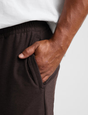 Close-up of a person wearing dark brown drawstring sweatpants with a side pocket, hand in pocket, paired with a white t-shirt against a light gray background.
