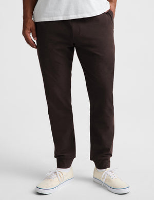 A person wearing dark brown jogger-style pants with elastic cuffs, paired with a white t-shirt and cream low-top sneakers, shown from the waist down on a light gray studio background.