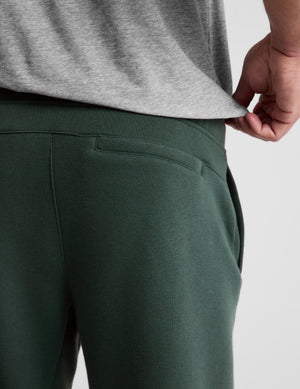 Close-up of a person wearing dark green sweatpants and a heather gray t-shirt, pulling the hem of the shirt to show the fit and waistband.