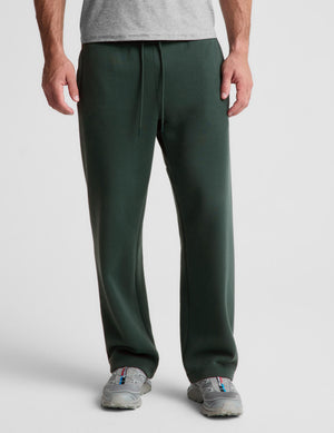 A person wearing dark green sweatpants with a drawstring waist, paired with a light gray t-shirt and gray sneakers, shown from the waist down on a light gray studio background.