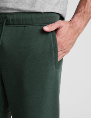Close-up of a person wearing dark green sweatpants with a side pocket and drawstring waistband, paired with a light gray heather top against a plain light gray background.
