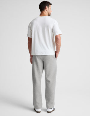 A man viewed from the back wearing a white short-sleeve t-shirt and light gray sweatpants against a light gray studio background.