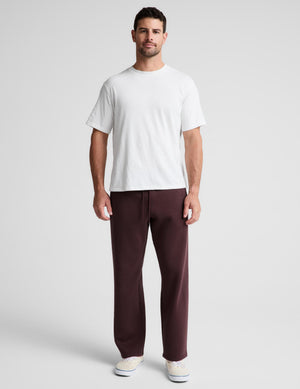 A man modeling a white crew-neck t-shirt and dark brown straight-leg sweatpants against a light gray studio background.