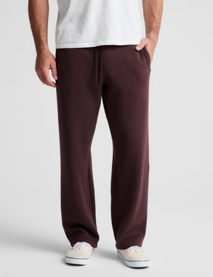 A person wearing dark burgundy sweatpants with a drawstring waist, paired with a white t-shirt and white sneakers, shown from the waist down on a light gray studio background.