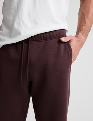 Close-up of a person wearing dark burgundy sweatpants with a drawstring waistband and side pocket, paired with a white t-shirt, against a light gray background.