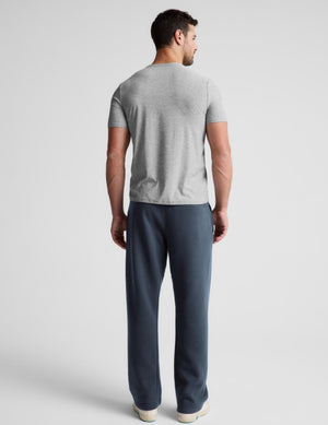 A man viewed from the back wearing a light gray short-sleeve t-shirt and dark slate blue pants against a light gray studio background.
