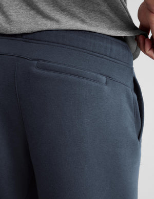 Close-up of a person wearing dark slate blue sweatpants, pulling the hem of a heather gray sweatshirt to show the elastic waistband and side pocket.