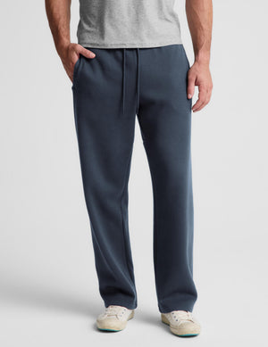A man wearing slate blue sweatpants with a drawstring waist, paired with a gray t-shirt and off-white sneakers, shown from the waist down on a light gray background.