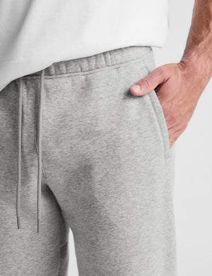 Close-up of a person wearing heather gray sweatpants with a drawstring waistband and side pocket, with one hand in the pocket against a light gray background.