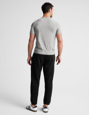 A man viewed from behind wearing a light gray short-sleeve t-shirt, black sweatpants, and white sneakers with black stripes, set against a plain light gray studio background.