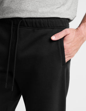 Close-up of a person wearing black sweatpants with a side pocket and drawstring waist, paired with a light gray heathered t-shirt. The person's hand is in the pocket, shown against a plain light gray background.