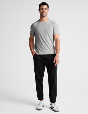 A man modeling a light gray short-sleeve crewneck t-shirt and black jogger pants with white sneakers, standing against a light gray studio background.