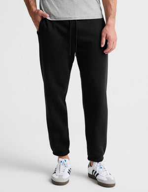 A person wearing black jogger sweatpants with a drawstring waist, paired with a light gray heathered t-shirt and white sneakers, shown from the waist down on a light gray studio background.