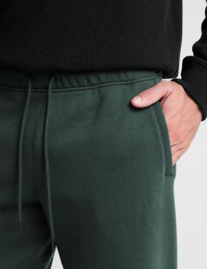 Close-up of a person wearing dark forest green sweatpants with a drawstring waist and side pocket, with a hand in the pocket and a black ribbed top visible.