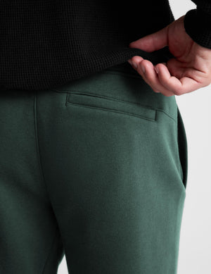 Close-up of a person wearing dark green pants and a black ribbed top, pulling the hem of the top to show the fabric and waistband construction.