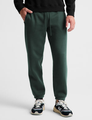A person wearing dark green jogger sweatpants with a drawstring waist, paired with a black sweatshirt and sneakers, shown from the chest down against a light gray studio background.