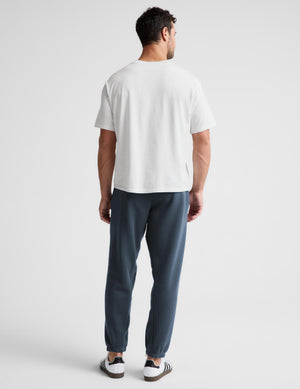 A man viewed from behind wearing a plain white short-sleeve t-shirt, slate blue jogger pants, and white sneakers with black stripes, set against a light gray studio background.