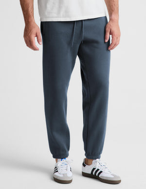 A person wearing dark slate blue jogger sweatpants with a white t-shirt and white sneakers, shown from the waist down against a light gray studio background.