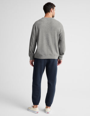A man viewed from behind wearing a light gray crewneck sweatshirt, navy blue jogger pants, and off-white slip-on sneakers against a light gray studio background.