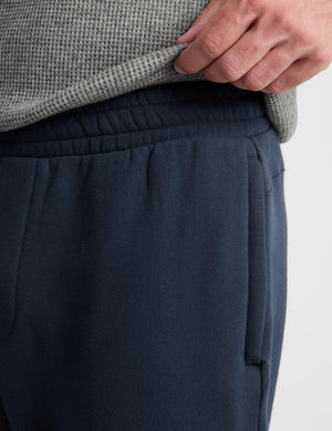 Close-up of a person wearing dark navy sweatpants and a gray textured knit top, with a hand pulling up the hem of the sweater to show the elastic waistband.