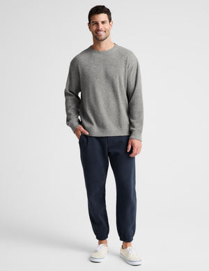 A man modeling a light gray crewneck sweater and dark navy jogger pants with white sneakers, standing against a light gray studio background.