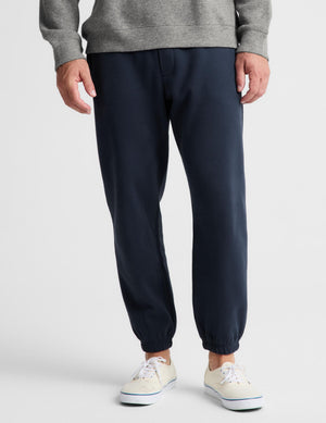 A person wearing dark navy jogger-style pants with elastic cuffs, paired with a gray ribbed sweater and off-white sneakers, shown from the waist down on a light gray studio background.