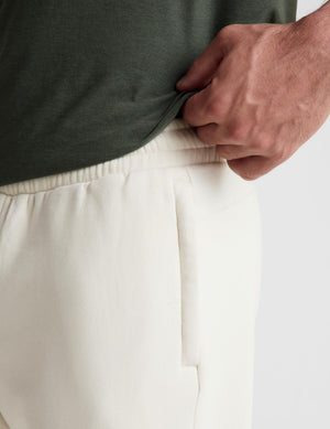 Close-up of a person wearing off-white drawstring pants and an olive green top, pulling the hem of the shirt to show the elastic waistband.