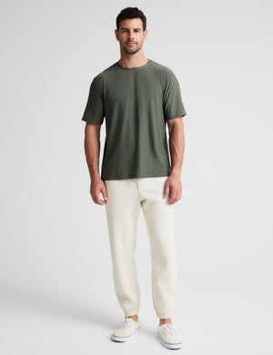 A man modeling a short-sleeve crewneck t-shirt in olive green, paired with cream-colored jogger pants and white sneakers, against a light gray studio background.