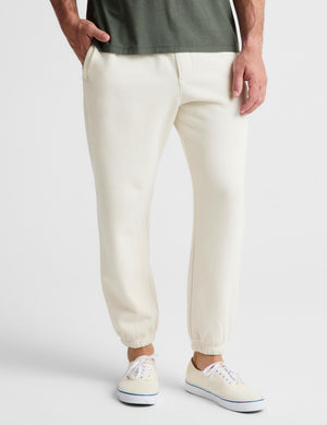 A person wearing cream-colored jogger pants with elastic cuffs, paired with a dark green t-shirt and cream low-top sneakers, shown from the waist down on a light gray studio background.