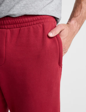 Close-up of a person wearing red fleece sweatpants with an elastic waistband and side pocket, with a hand in the pocket and a gray heather t-shirt partially visible.