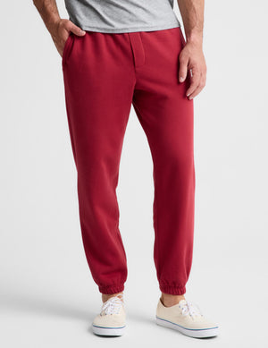 A person wearing red jogger-style pants with elastic cuffs, paired with a light gray t-shirt and cream sneakers, shown from the waist down on a light gray studio background.