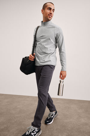 men's model in gray long-sleeve shirt and dark pants holding a black bag and silver water bottle against a plain background. 