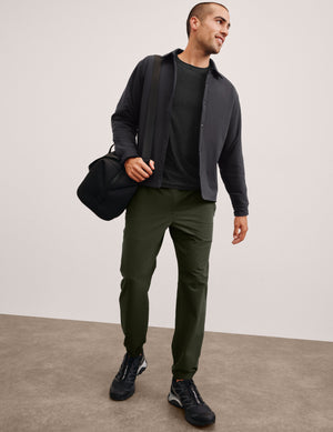 man holding a black duffel bag wearing a dark jacket, black shirt, and green chino pants on a plain background. 