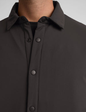 Man wearing a dark brown jacket over a black shirt with green pants on a light gray background. 