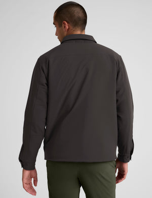 Man wearing a dark brown jacket over a black shirt with green pants on a light gray background. 