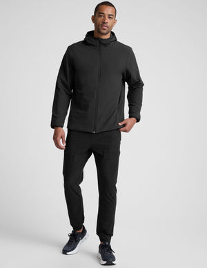 male model wearing a black zip-up jacket with a hood and pockets against a plain background. 