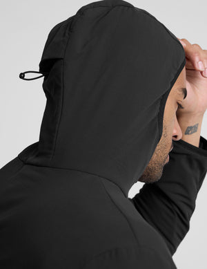 male model wearing a black zip-up jacket with a hood and pockets against a plain background. 
