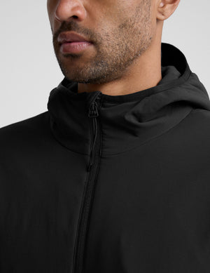 male model wearing a black zip-up jacket with a hood and pockets against a plain background. 