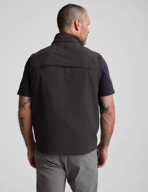 Man wearing a black vest over a dark shirt with a neutral background. 