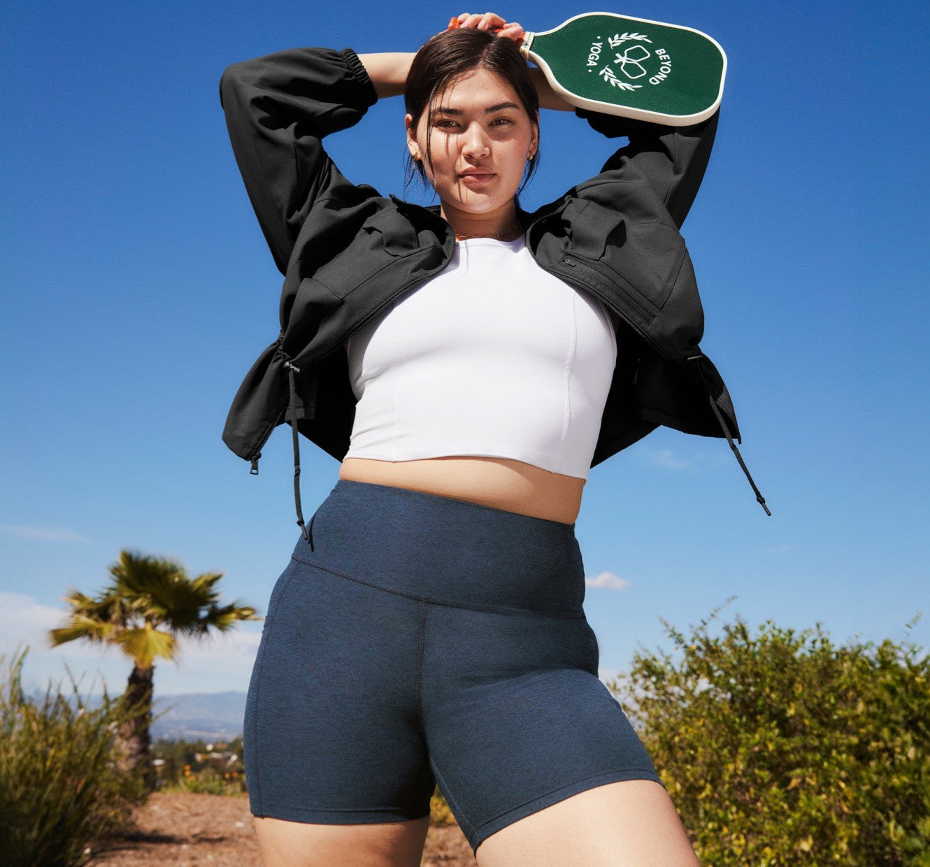 model is wearing a white high-neck cropped tank top and blue high-waisted biker shorts. 
