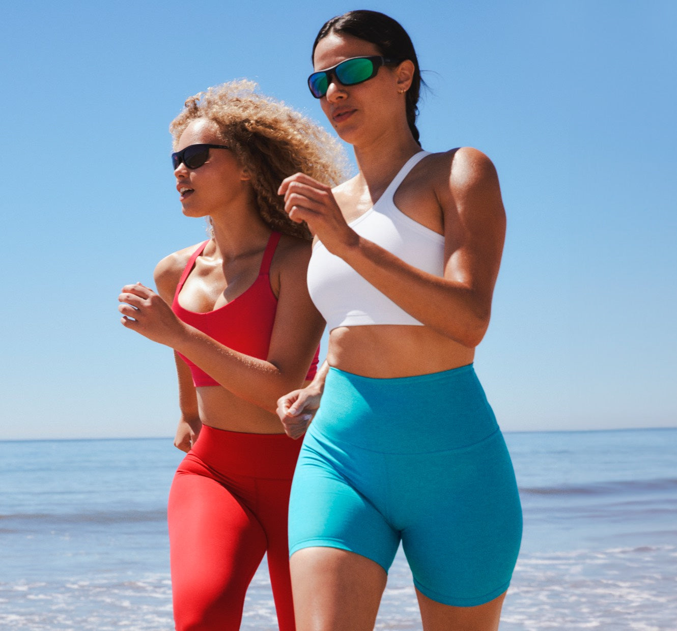 model is wearing a red racerback bra and red high-waisted midi leggings. model on the right is wearing a white one-shoulder bra top and blue high-waisted biker shorts. 