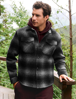 A man wearing a dark gray and black plaid button-up fleece jacket over a burgundy hoodie, standing outdoors on a wooden deck with trees in the background.